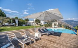 Modern outdoor pool area with lounge chairs and a large umbrella, surrounded by lush greenery and scenic mountain views, ideal for relaxation and leisure.
