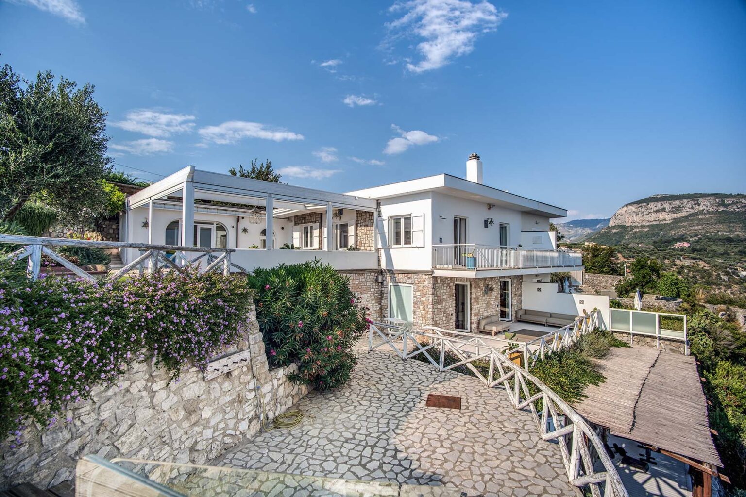 Modern hillside villa with stone exterior, surrounded by greenery and featuring a spacious terrace, under a clear blue sky. | Only on SopranoVillas