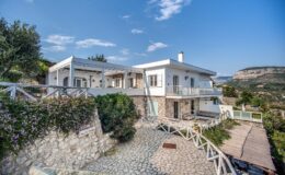Modern hillside villa with stone exterior, surrounded by greenery and featuring a spacious terrace, under a clear blue sky.