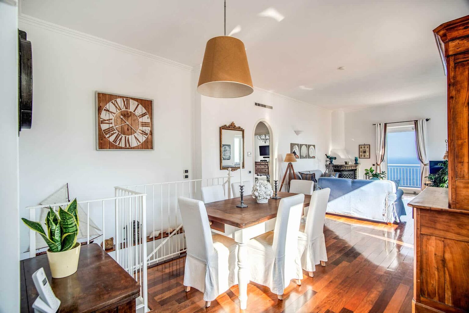 Bright and airy interior of a modern dining area featuring a wooden dining table with white chairs, a large clock on the wall, and a stylish lamp. The space is complemented by a potted plant and an open layout leading to a cozy living area with natural light streaming in. | Only on SopranoVillas