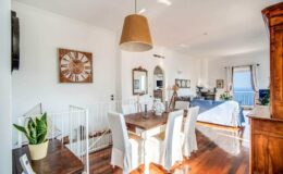Bright and airy interior of a modern dining area featuring a wooden dining table with white chairs, a large clock on the wall, and a stylish lamp. The space is complemented by a potted plant and an open layout leading to a cozy living area with natural light streaming in.