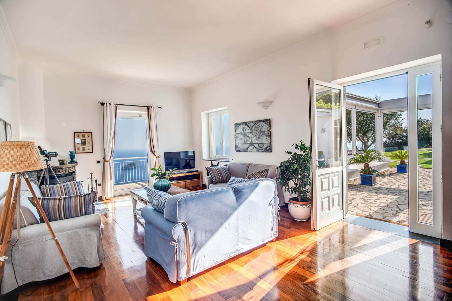 Bright and airy living room with large windows showcasing a scenic view, featuring a comfortable blue sofa, wooden flooring, and a cozy atmosphere. The room opens to an outdoor patio, enhancing the connection to nature. | Only on SopranoVillas