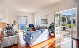 Bright and airy living room with large windows showcasing a scenic view, featuring a comfortable blue sofa, wooden flooring, and a cozy atmosphere. The room opens to an outdoor patio, enhancing the connection to nature.