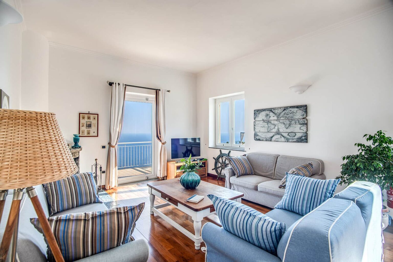 Bright and airy living room featuring a cozy seating area with striped and solid sofas, a wooden coffee table, and a television. Large windows offer a view of the ocean, complemented by light curtains and indoor plants for a refreshing atmosphere. | Only on SopranoVillas