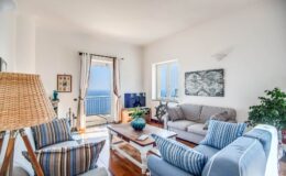 Bright and airy living room featuring a cozy seating area with striped and solid sofas, a wooden coffee table, and a television. Large windows offer a view of the ocean, complemented by light curtains and indoor plants for a refreshing atmosphere.