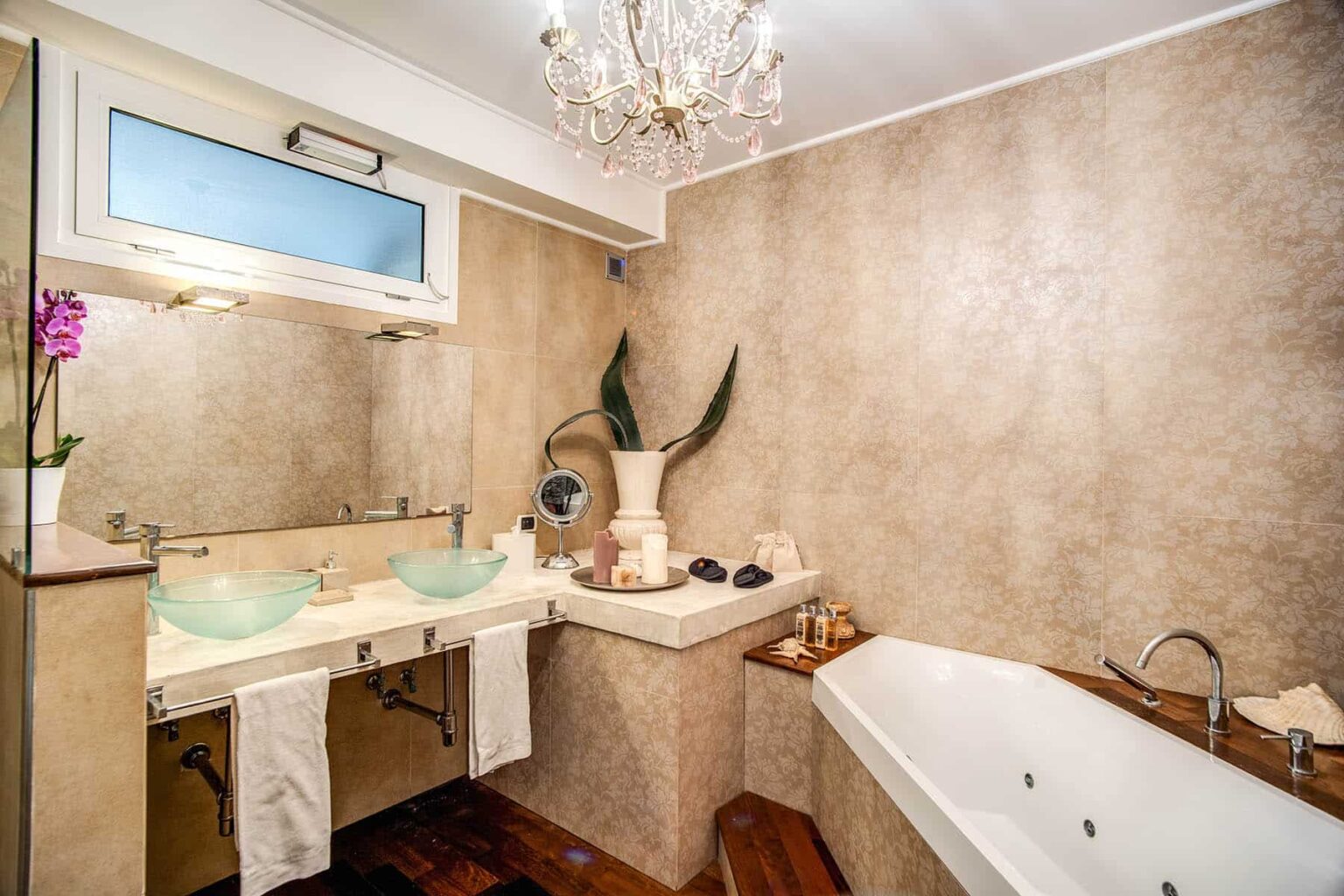 Elegant modern bathroom featuring a luxurious bathtub, dual glass sinks, and stylish decor with a chandelier and potted plants. | Only on SopranoVillas