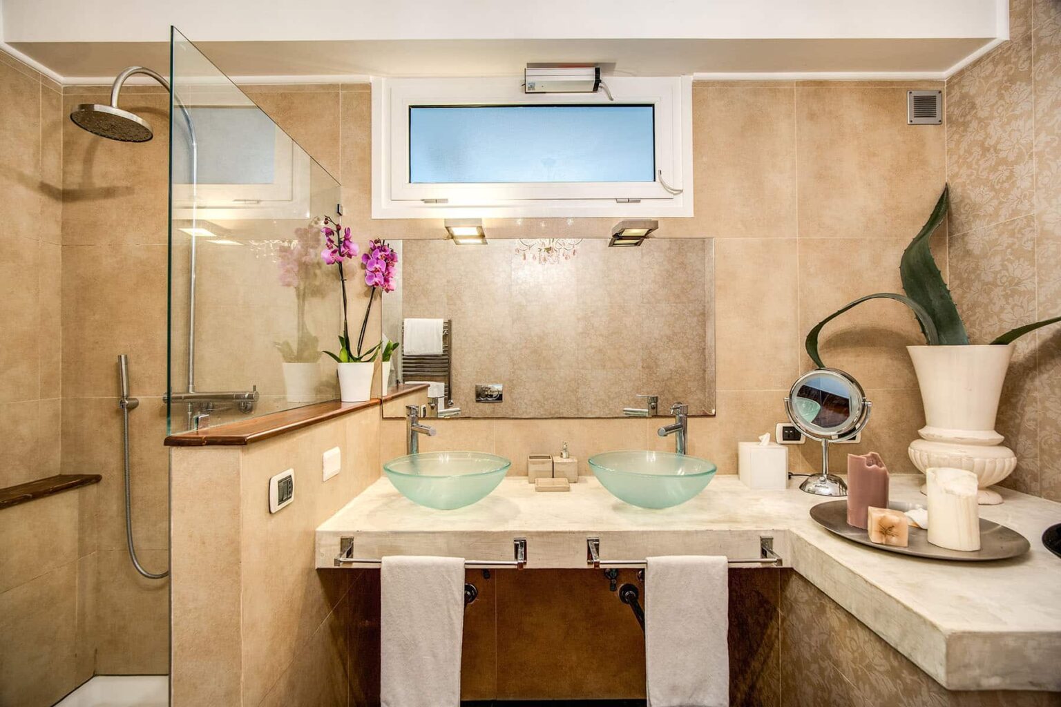 Modern bathroom featuring dual glass vessel sinks, a spacious shower with glass enclosure, elegant beige stone walls, and decorative elements like potted orchids and a stylish vanity. | Only on SopranoVillas