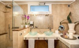 Modern bathroom featuring dual glass vessel sinks, a spacious shower with glass enclosure, elegant beige stone walls, and decorative elements like potted orchids and a stylish vanity.