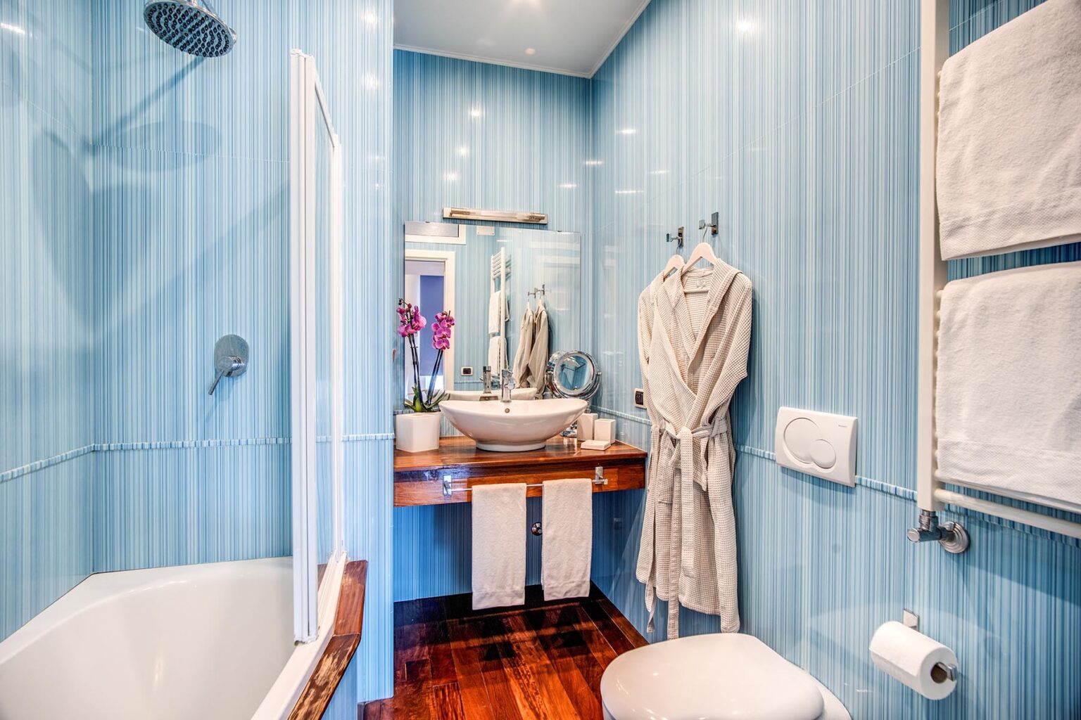 Modern bathroom featuring light blue striped walls, a bathtub, a stylish sink with a mirror, and elegant decor, including a robe and towels. | Only on SopranoVillas