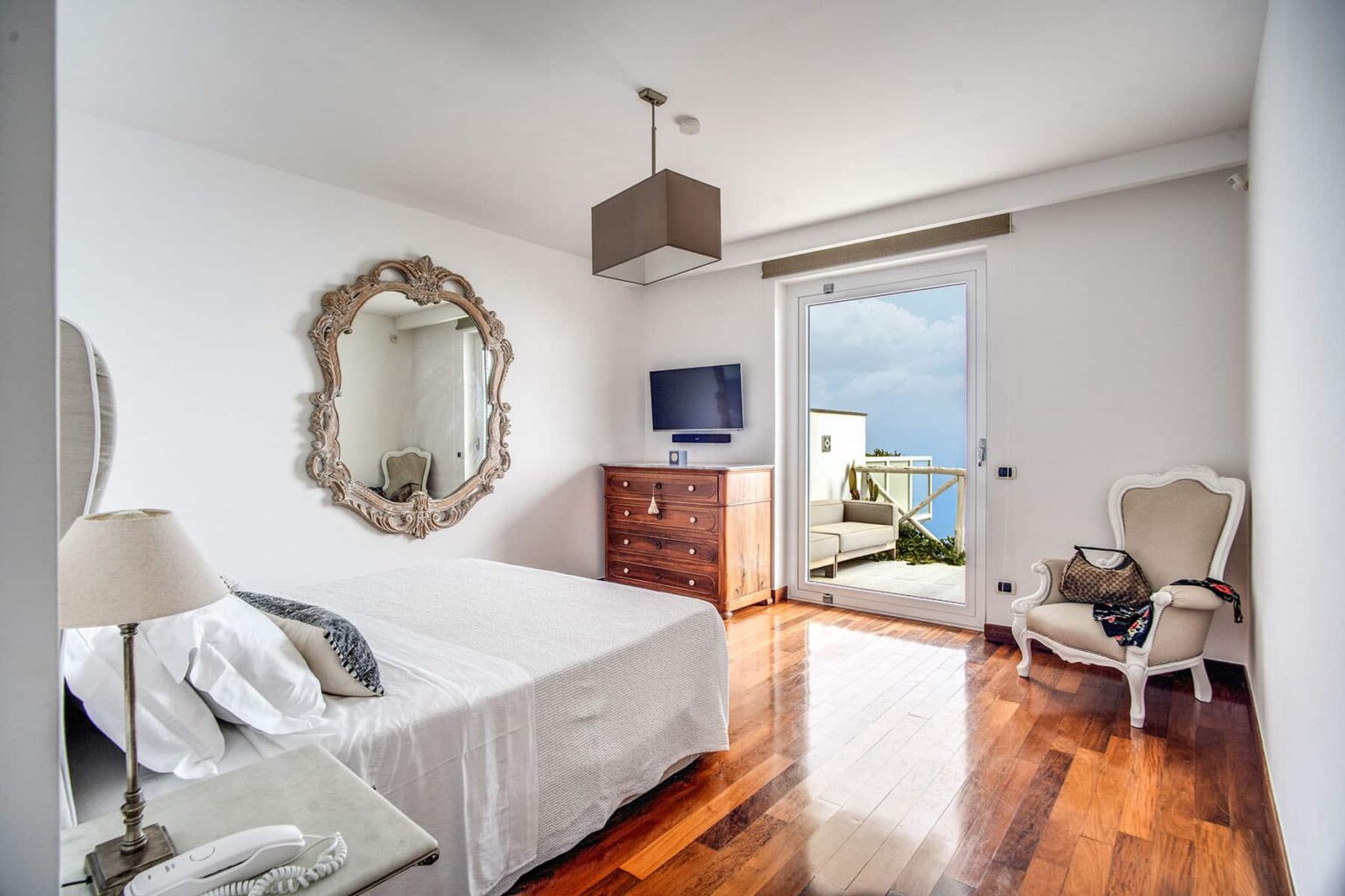 Modern bedroom featuring a white bed, elegant mirror, wooden dresser, and a stylish armchair, with large windows offering a view and natural light. | Only on SopranoVillas