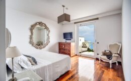 Modern bedroom featuring a white bed, elegant mirror, wooden dresser, and a stylish armchair, with large windows offering a view and natural light.