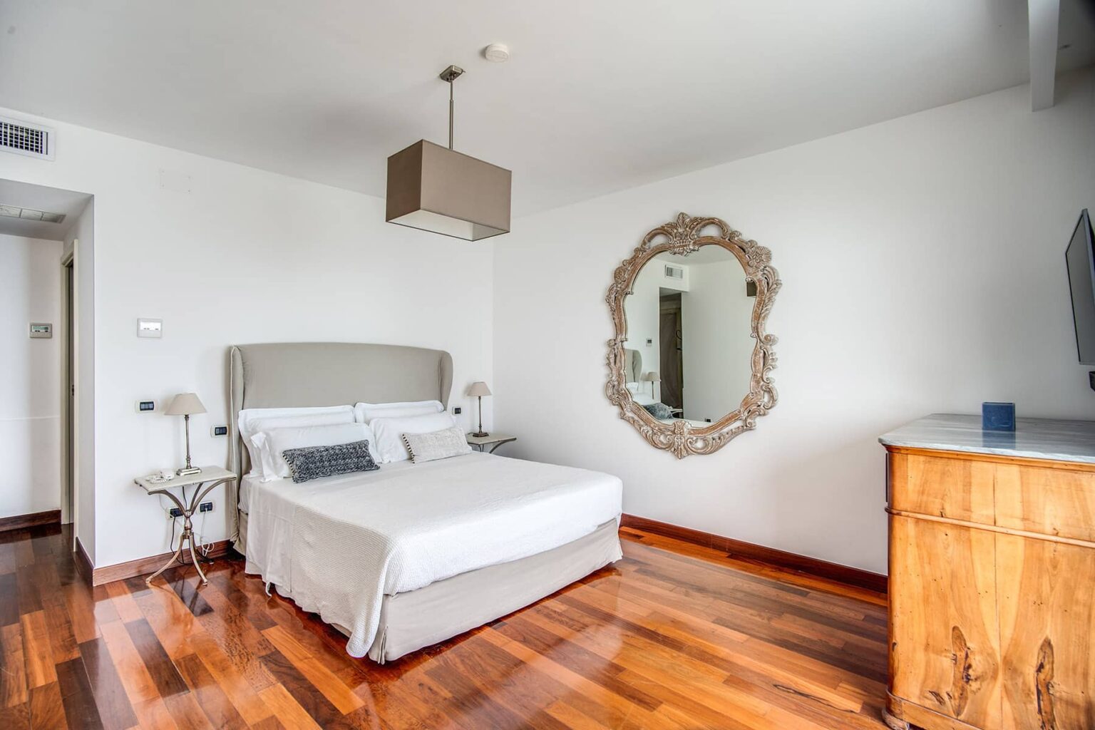 Modern bedroom featuring a queen-sized bed with a light gray upholstered headboard, elegant wooden flooring, a stylish mirror, and a contemporary ceiling light fixture, creating a serene and inviting atmosphere. | Only on SopranoVillas
