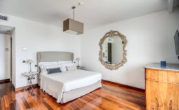 Modern bedroom featuring a queen-sized bed with a light gray upholstered headboard, elegant wooden flooring, a stylish mirror, and a contemporary ceiling light fixture, creating a serene and inviting atmosphere.