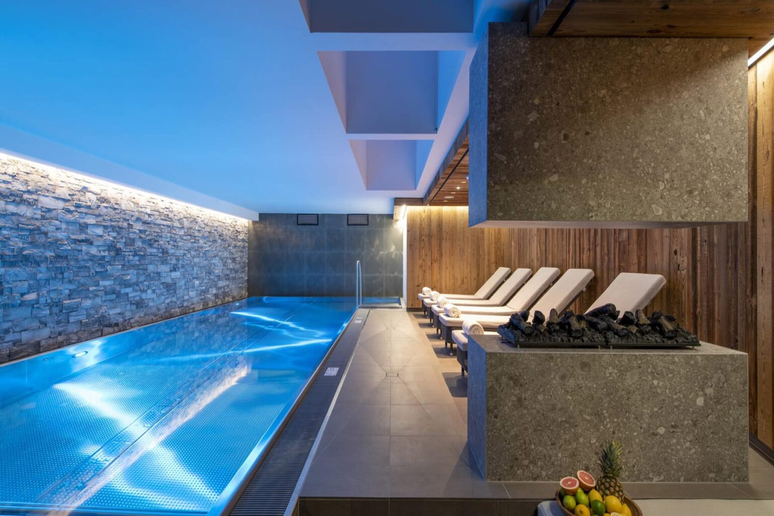 Modern indoor swimming pool area featuring sleek stone and wood accents, lounge chairs, and ambient lighting for a relaxing atmosphere. | Only on SopranoVillas