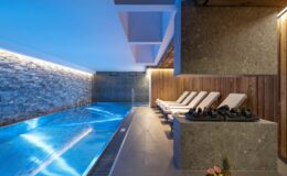 Modern indoor swimming pool area featuring sleek stone and wood accents, lounge chairs, and ambient lighting for a relaxing atmosphere.
