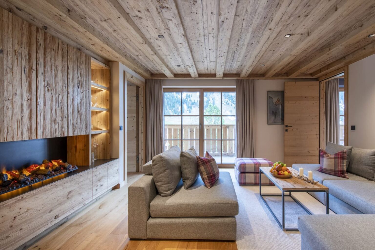 Cozy modern living room with wooden ceiling and walls, featuring a stylish fireplace, comfortable sofas with pillows, and a coffee table adorned with fruit. Large windows provide natural light and a view of the outdoors. | Only on SopranoVillas