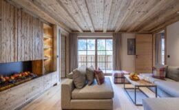 Cozy modern living room with wooden ceiling and walls, featuring a stylish fireplace, comfortable sofas with pillows, and a coffee table adorned with fruit. Large windows provide natural light and a view of the outdoors.