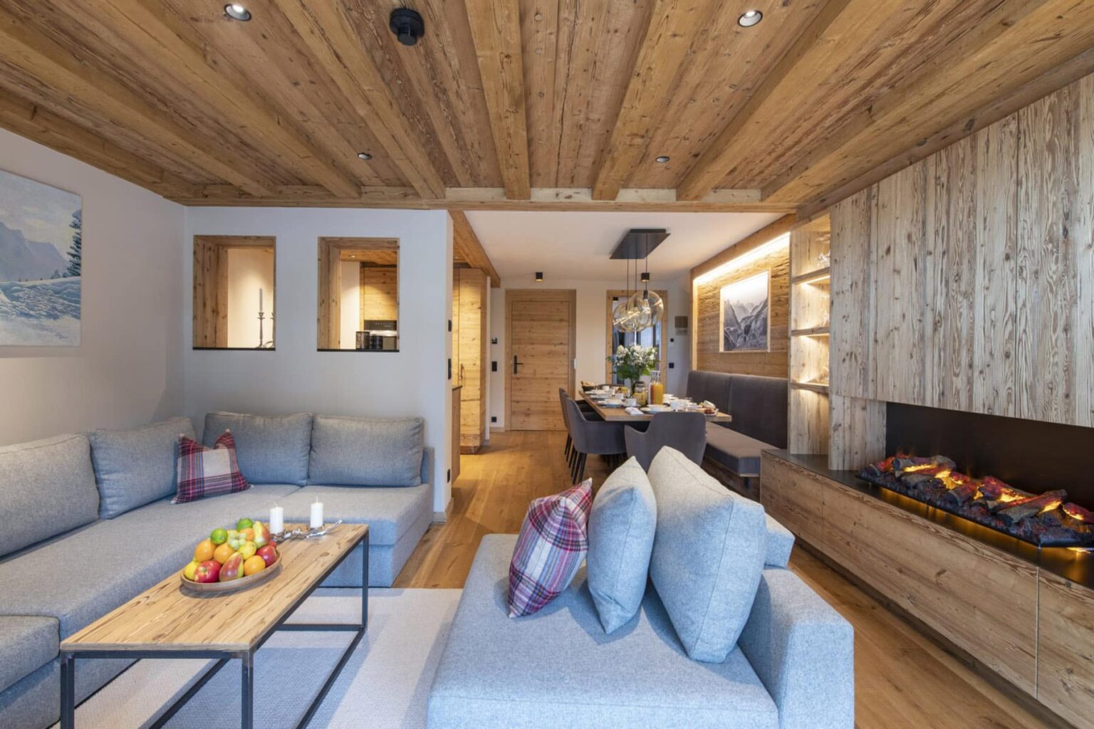 Modern living room interior featuring a cozy sofa, wooden ceiling beams, and a stylish fireplace. The space is decorated with natural materials, soft cushions, and a fruit centerpiece on the coffee table, creating a warm and inviting atmosphere. | Only on SopranoVillas