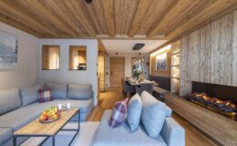 Modern living room interior featuring a cozy sofa, wooden ceiling beams, and a stylish fireplace. The space is decorated with natural materials, soft cushions, and a fruit centerpiece on the coffee table, creating a warm and inviting atmosphere.
