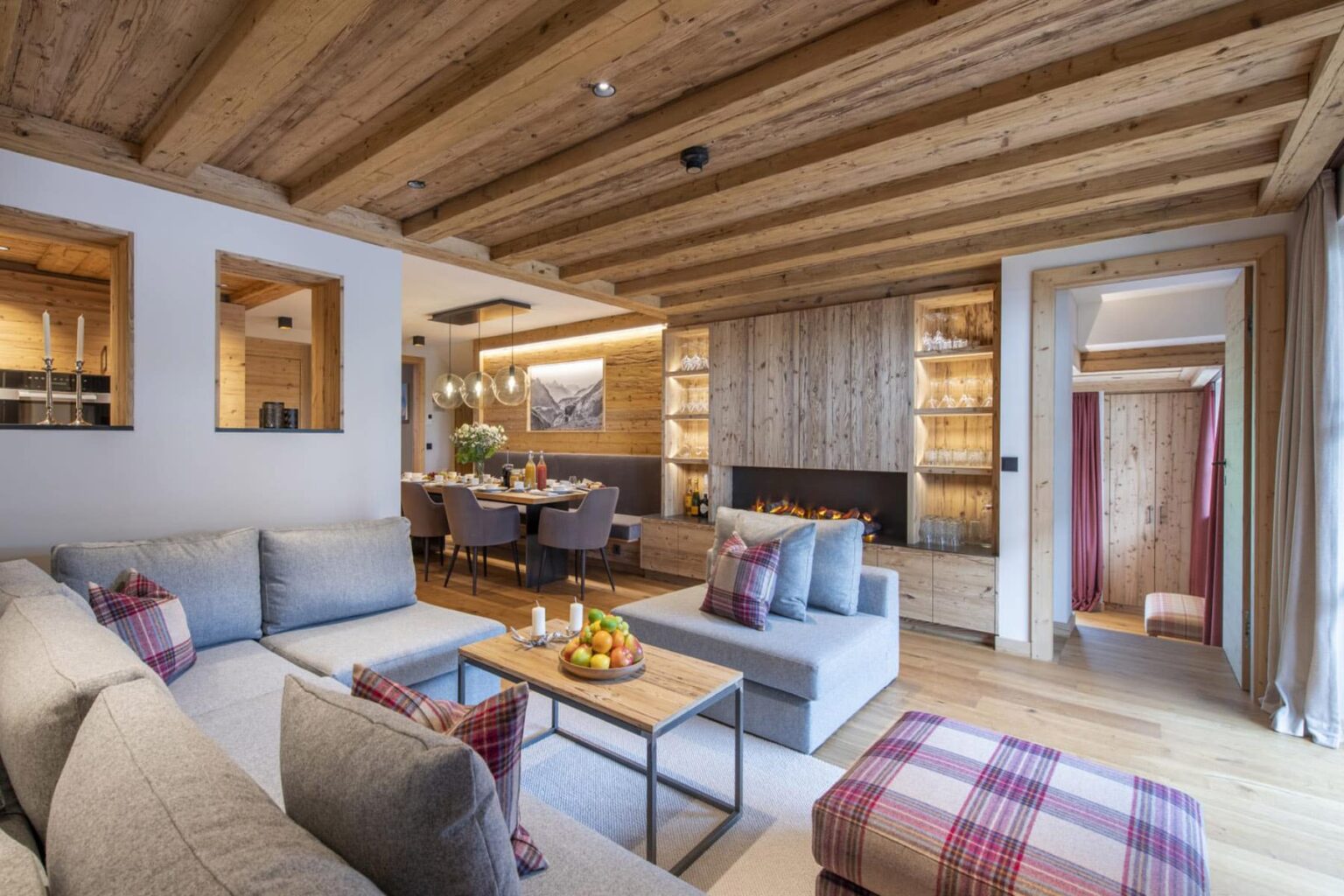 Modern living room with wooden beams, stylish gray sofas, a coffee table with fruit, and a cozy atmosphere, leading to a dining area and kitchen in a contemporary home. | Only on SopranoVillas