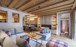Modern living room with wooden beams, stylish gray sofas, a coffee table with fruit, and a cozy atmosphere, leading to a dining area and kitchen in a contemporary home.