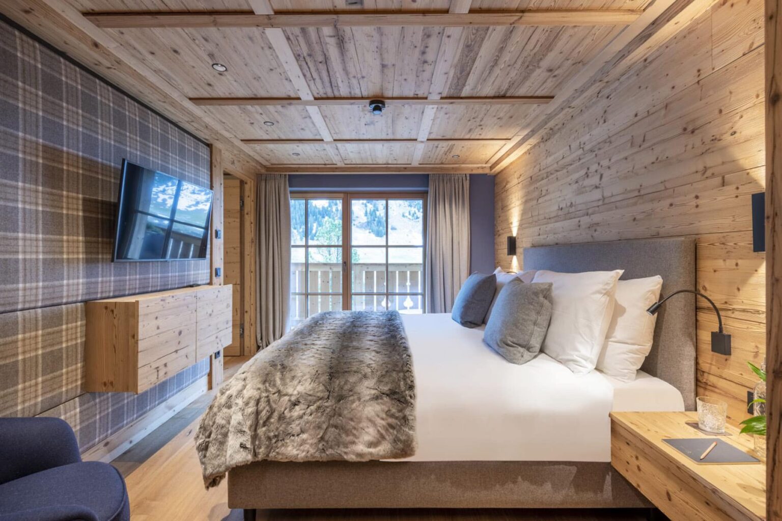 Cozy hotel bedroom featuring wooden walls, a plush bed with decorative pillows, a large window with natural light, a flat-screen TV, and stylish furnishings, perfect for a relaxing stay. | Only on SopranoVillas