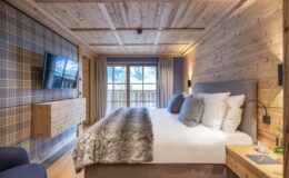 Cozy hotel bedroom featuring wooden walls, a plush bed with decorative pillows, a large window with natural light, a flat-screen TV, and stylish furnishings, perfect for a relaxing stay.