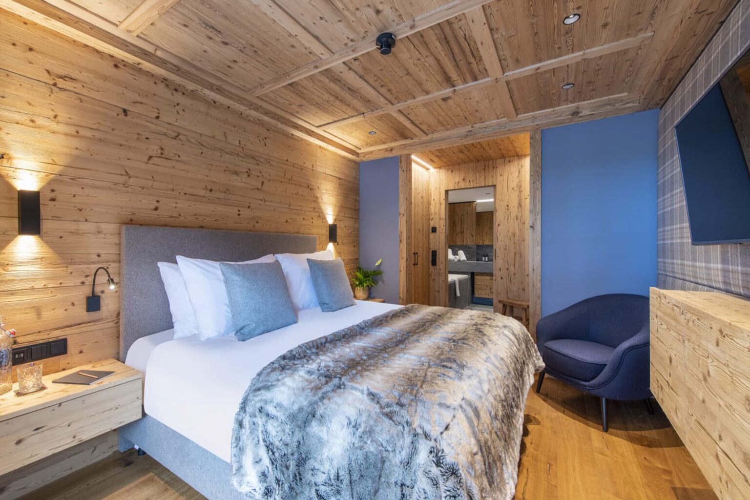 Cozy hotel room featuring a wooden interior, a plush king-sized bed with decorative pillows, a stylish chair, and modern amenities, perfect for a relaxing stay. | Only on SopranoVillas