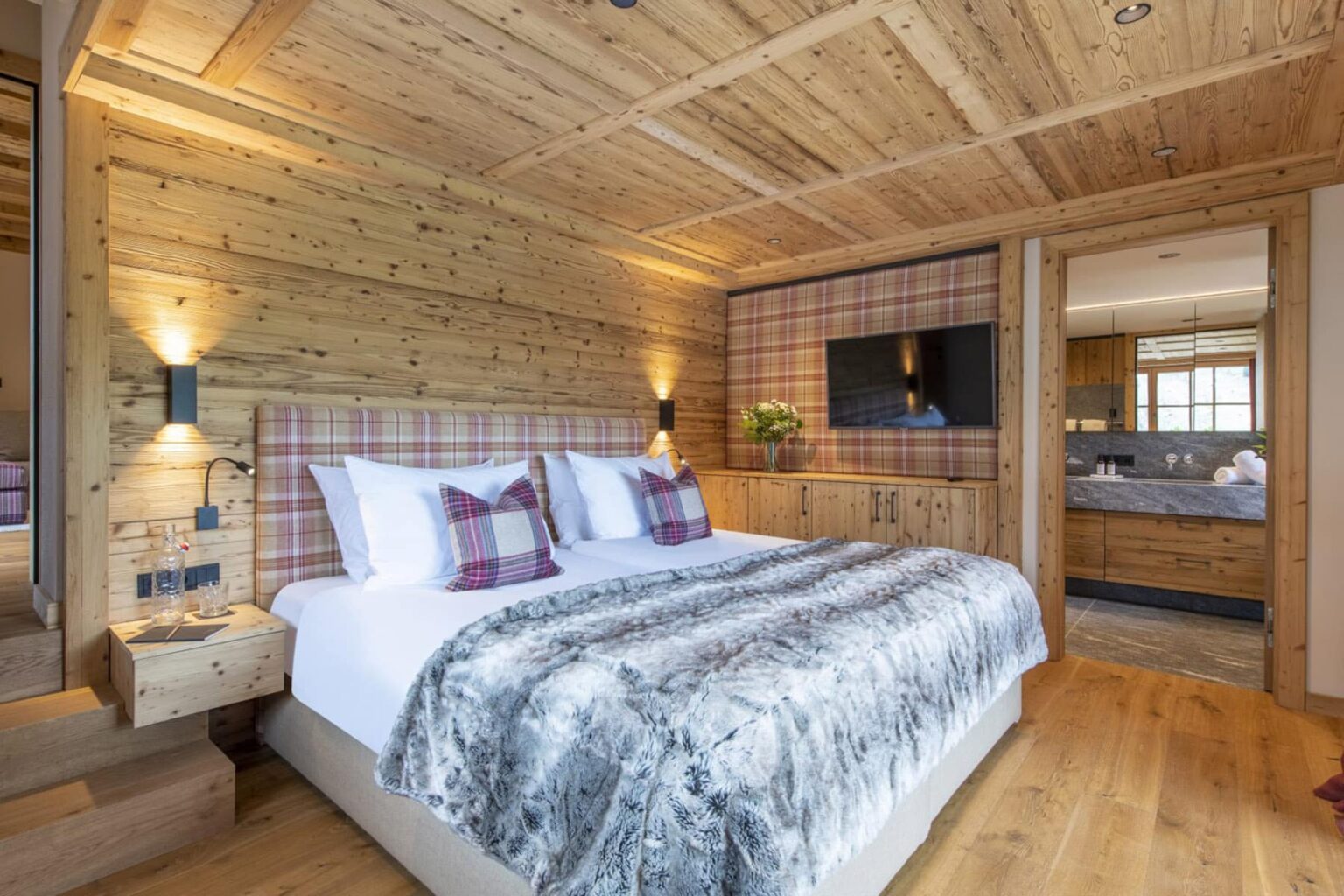Cozy bedroom with wooden walls, featuring a king-size bed with a plush gray blanket, decorative plaid pillows, and warm lighting. The room includes a flat-screen TV and connects to a stylish bathroom, creating a rustic yet modern retreat. | Only on SopranoVillas