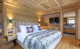 Cozy bedroom with wooden walls, featuring a king-size bed with a plush gray blanket, decorative plaid pillows, and warm lighting. The room includes a flat-screen TV and connects to a stylish bathroom, creating a rustic yet modern retreat.