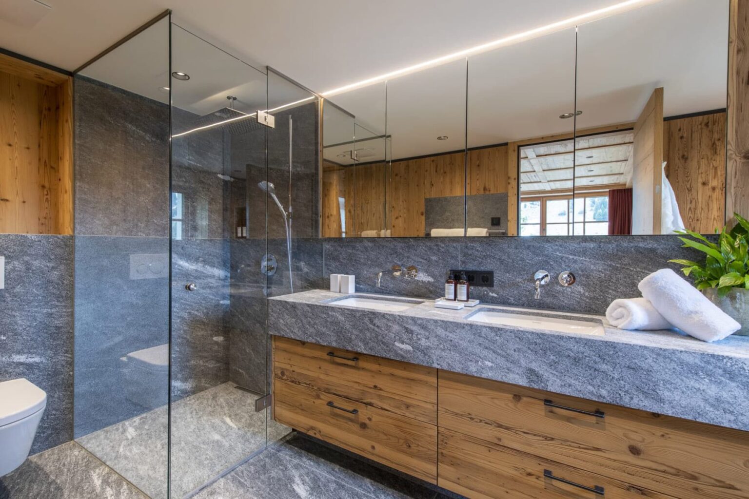 Modern bathroom featuring a sleek glass shower, wooden cabinetry, and marble countertops, complemented by natural light and greenery for a contemporary aesthetic. | Only on SopranoVillas