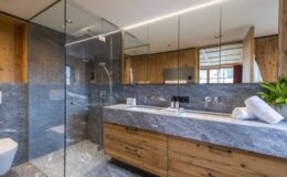 Modern bathroom featuring a sleek glass shower, wooden cabinetry, and marble countertops, complemented by natural light and greenery for a contemporary aesthetic.