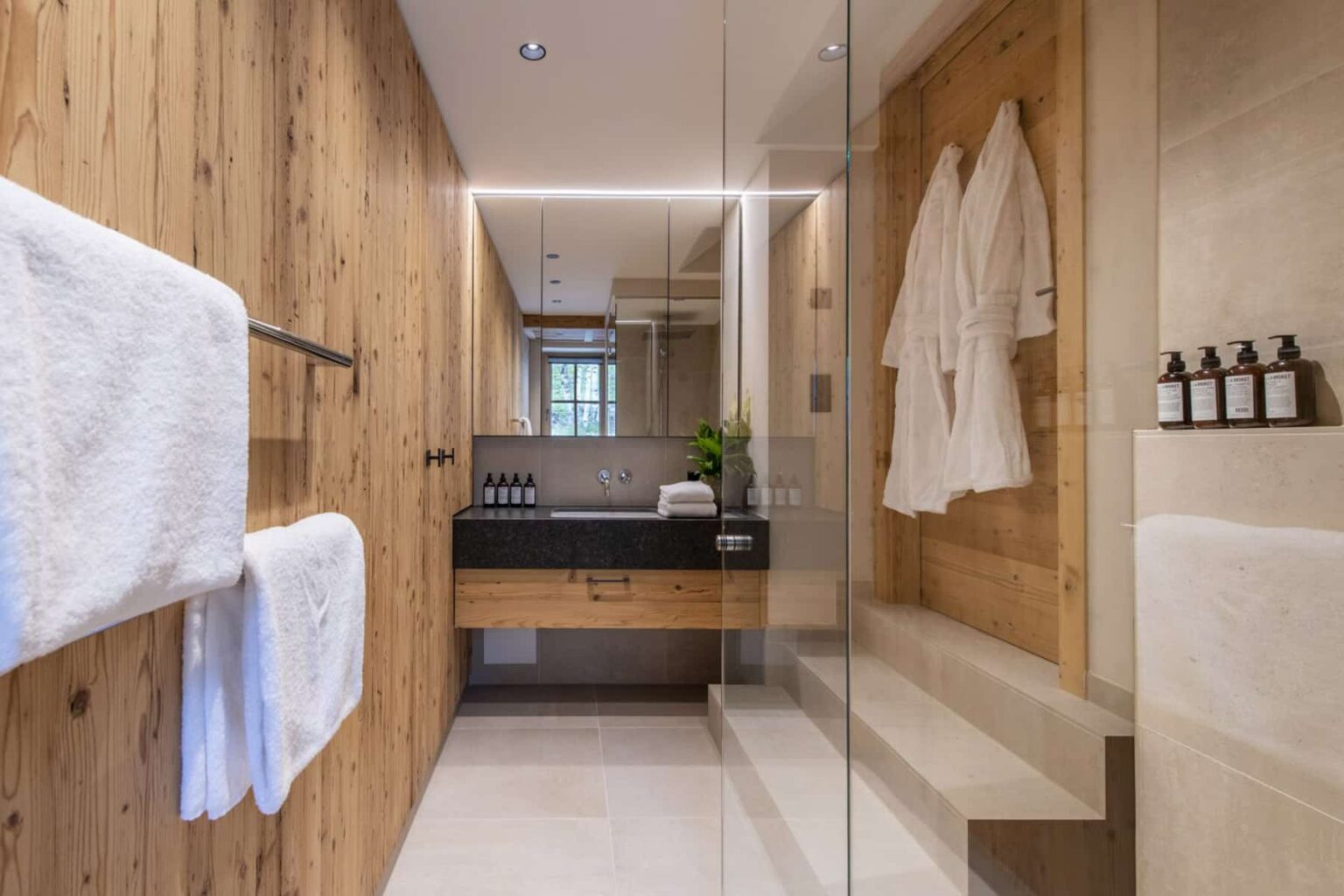 Modern bathroom design featuring wooden accents, a glass shower enclosure, plush white towels, and elegant toiletries. | Only on SopranoVillas