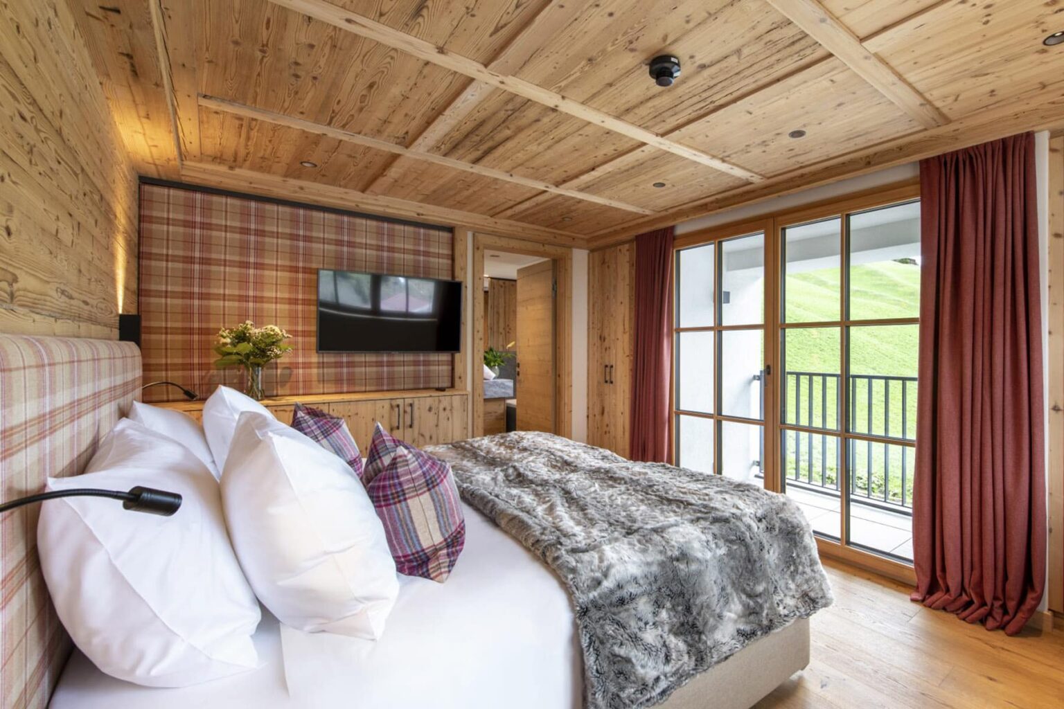 Cozy bedroom interior featuring wooden walls, plush bedding with decorative pillows, a flat-screen TV, and large windows with views of nature, designed for comfort and relaxation. | Only on SopranoVillas