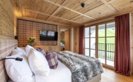 Cozy bedroom interior featuring wooden walls, plush bedding with decorative pillows, a flat-screen TV, and large windows with views of nature, designed for comfort and relaxation.