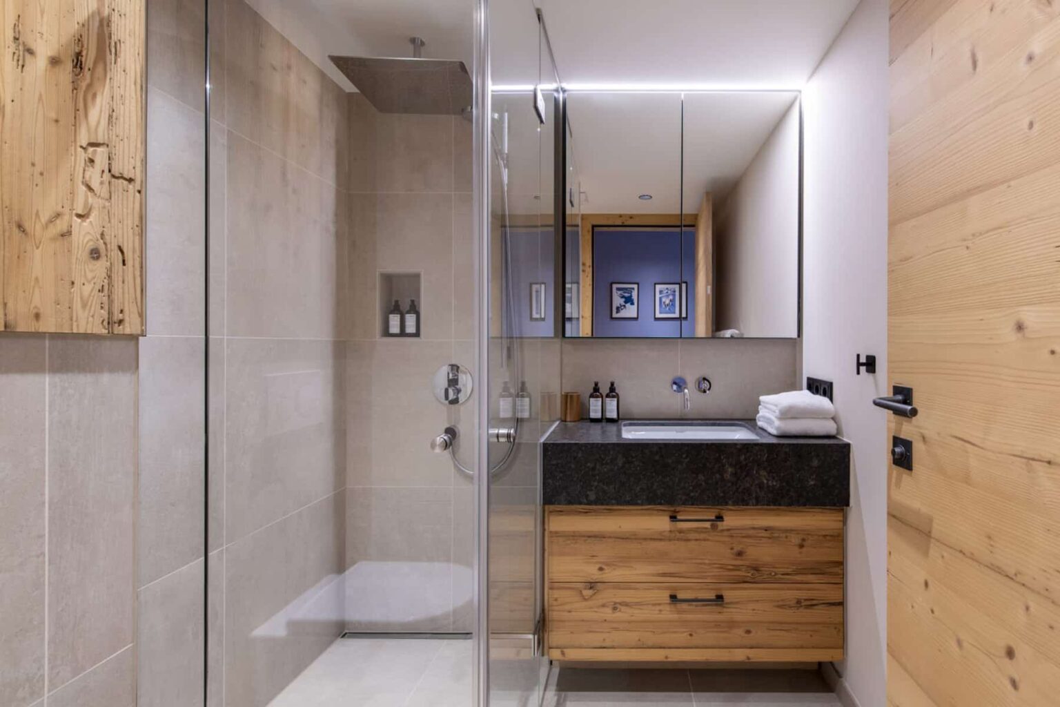 Modern bathroom interior featuring a glass shower, wooden accents, and a sleek black countertop with toiletries. Natural light enhances the spacious and stylish design. | Only on SopranoVillas