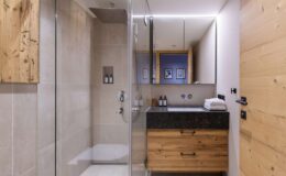 Modern bathroom interior featuring a glass shower, wooden accents, and a sleek black countertop with toiletries. Natural light enhances the spacious and stylish design.