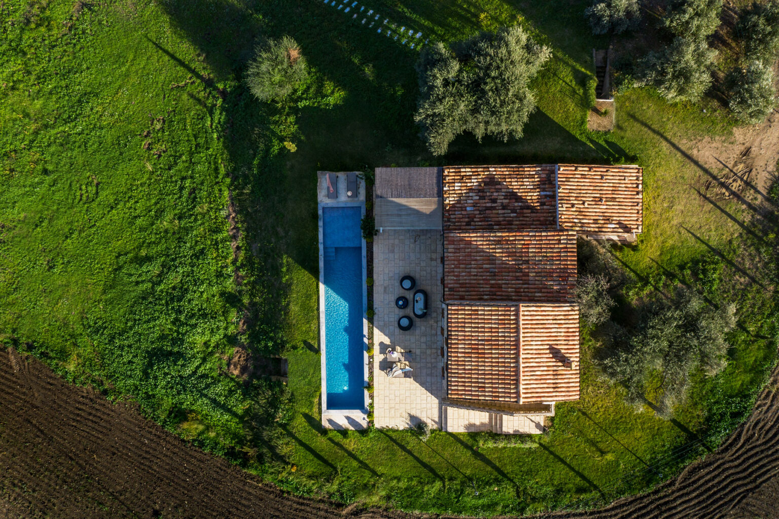 Aerial view of a modern villa with a swimming pool surrounded by lush green fields and olive trees, showcasing outdoor seating and a rustic tiled roof. | Only on SopranoVillas