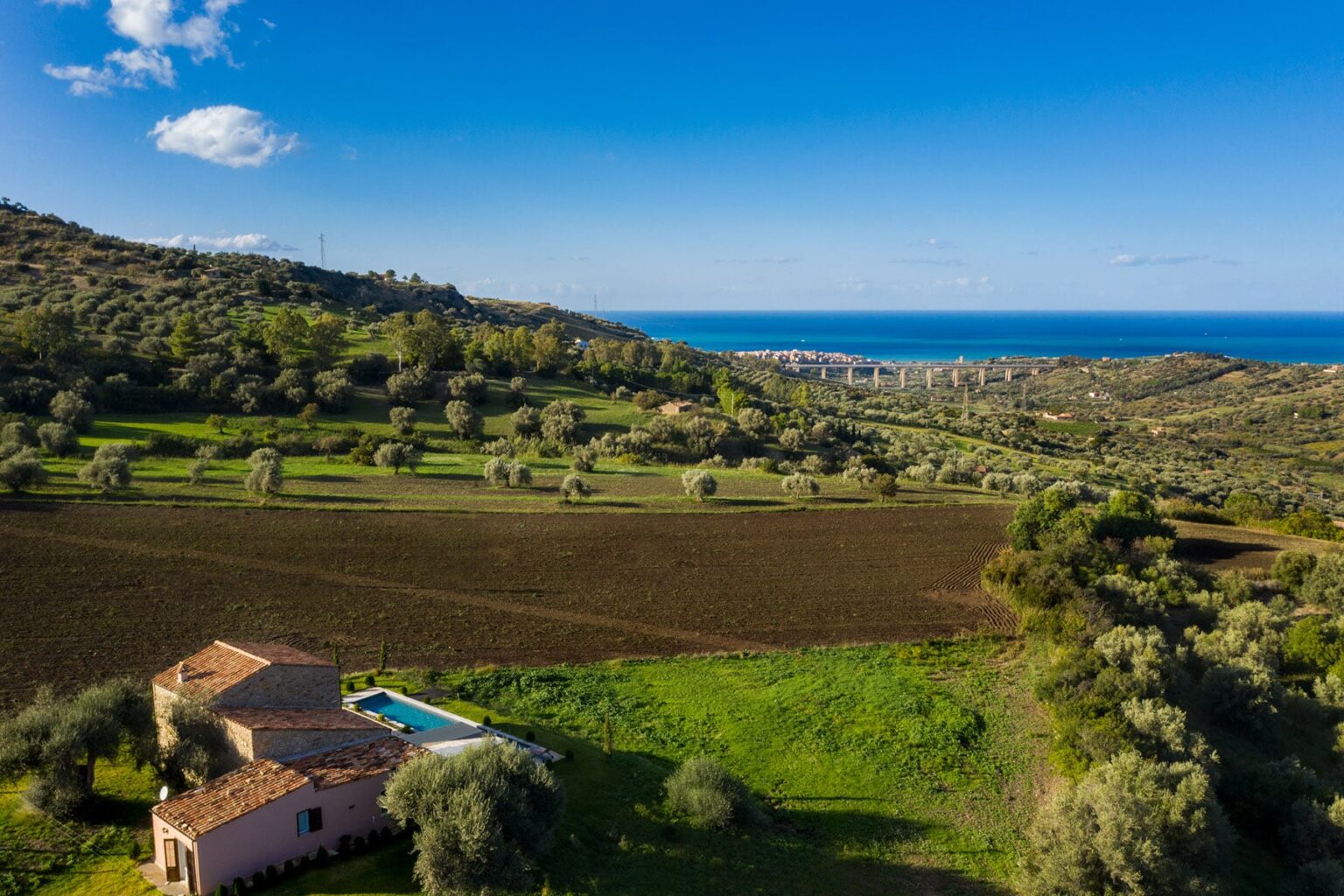 Aerial view of a scenic landscape featuring rolling hills, olive trees, and a tranquil sea in the background, highlighting a rustic villa with a pool nestled amidst the greenery. | Only on SopranoVillas