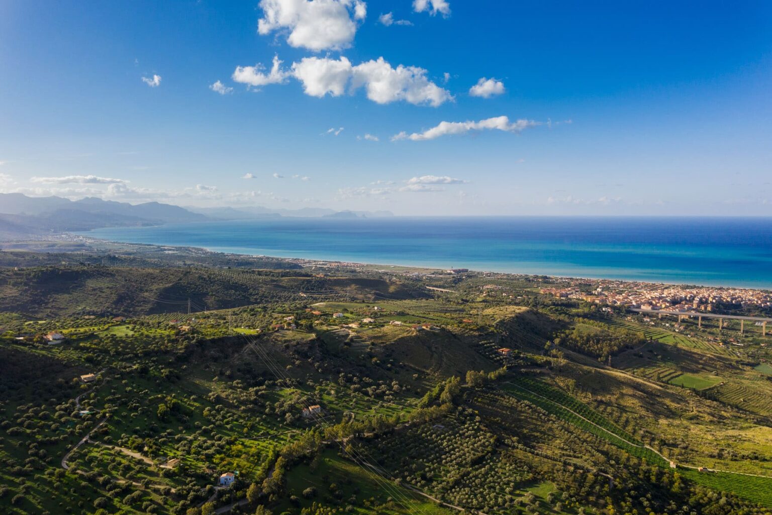 Aerial view of a coastal landscape featuring lush green hills and a clear blue sea, with a distant shoreline and mountains under a bright sky. | Only on SopranoVillas