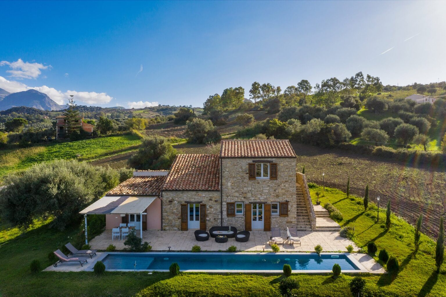 Aerial view of a charming stone villa with a swimming pool, surrounded by lush greenery and rolling hills, set against a clear blue sky. Ideal for vacation rentals or countryside retreats. | Only on SopranoVillas