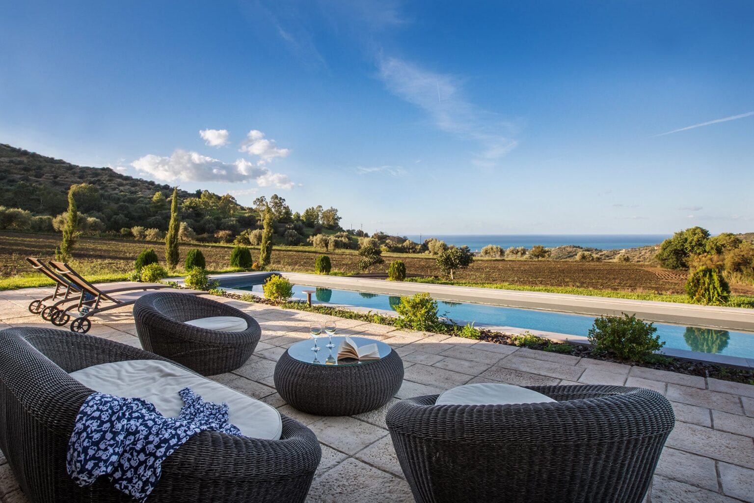 Modern outdoor patio with wicker chairs and a glass table overlooking a serene pool and scenic landscape. Lush greenery and distant hills under a clear blue sky enhance the tranquil atmosphere. Ideal for relaxation and outdoor gatherings. | Only on SopranoVillas