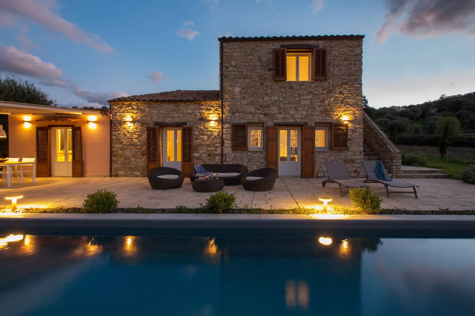 Stone villa at dusk with illuminated outdoor seating area and swimming pool, surrounded by lush greenery in a tranquil setting. | Only on SopranoVillas