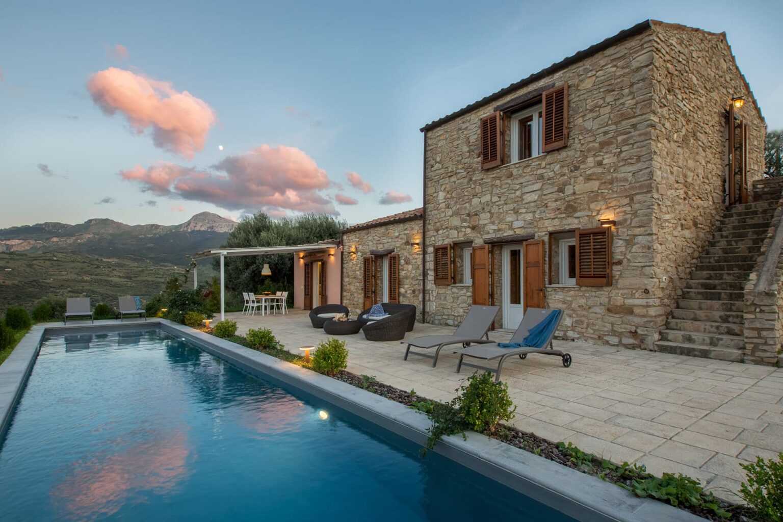 Stone villa with a swimming pool, surrounded by mountains and a sunset sky, featuring outdoor seating and a landscaped patio. Ideal for vacation rentals or luxury getaways. | Only on SopranoVillas