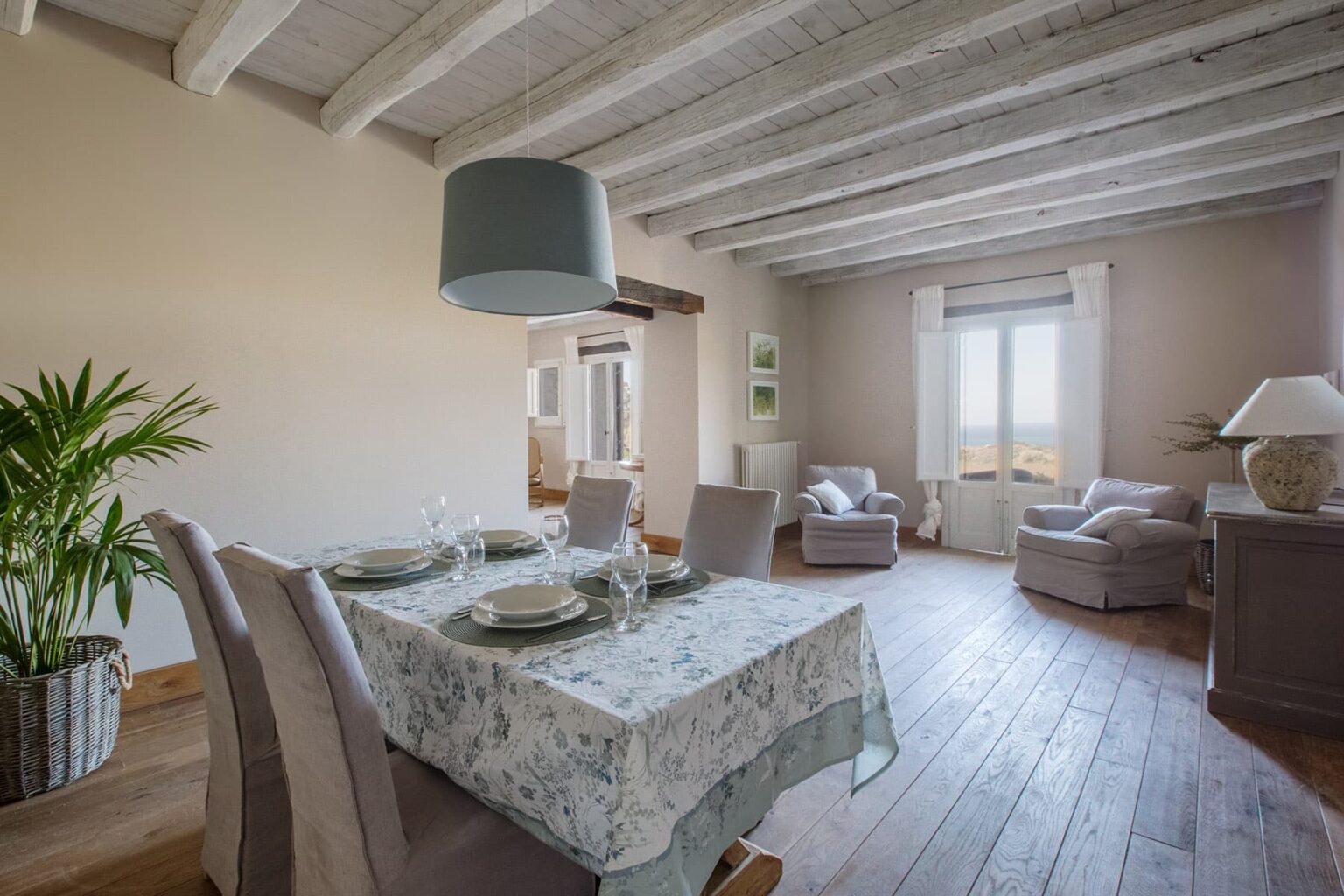 Modern dining room with a wooden floor, featuring a floral tablecloth, set dining table, and cozy seating area with armchairs. Natural light streams through large windows, enhancing the inviting atmosphere. | Only on SopranoVillas