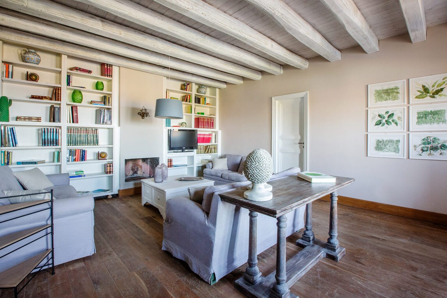 Modern living room featuring a cozy seating area with gray sofas, a wooden coffee table, and a stylish bookshelf filled with books and decorative items. The space is illuminated by natural light, highlighting the wooden floor and framed botanical art on the walls. | Only on SopranoVillas