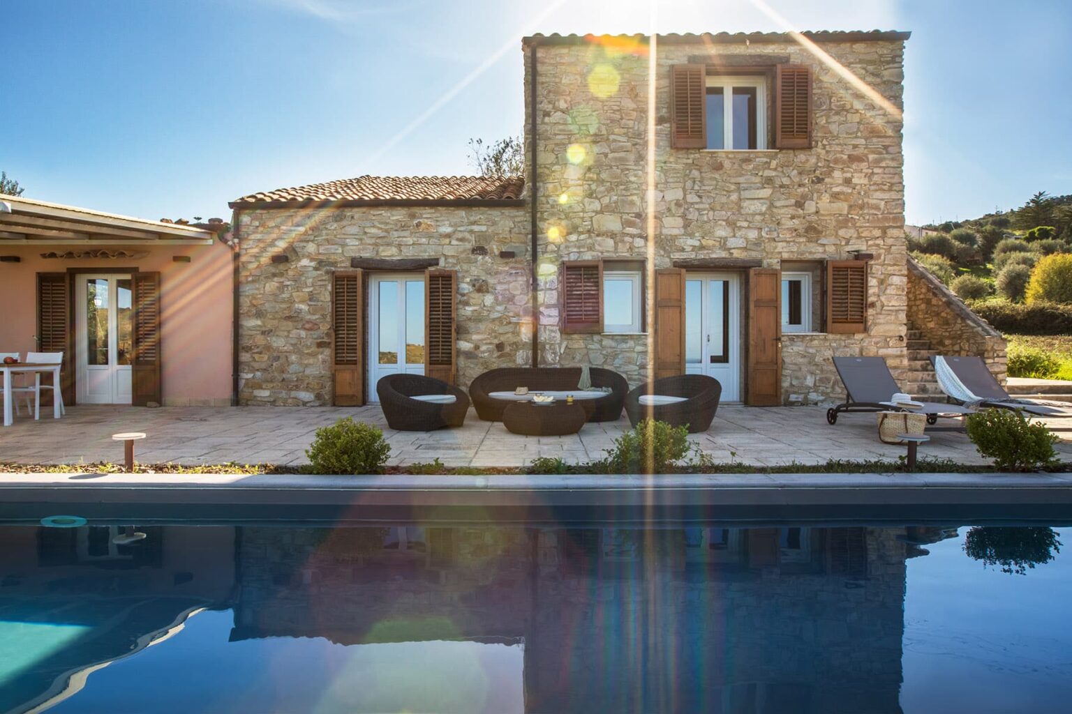 Stone villa with wooden shutters and outdoor seating area, featuring a swimming pool and sunny landscape. Ideal for vacation rentals and relaxing getaways. | Only on SopranoVillas