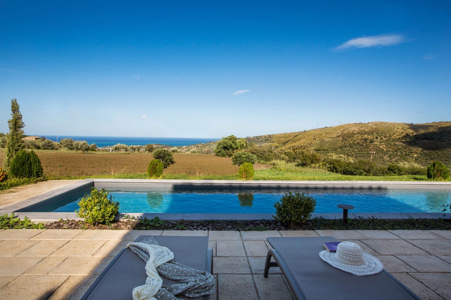 Scenic view of a luxurious infinity pool overlooking rolling hills and the ocean, with sun loungers and a sun hat in the foreground, ideal for a relaxing getaway. | Only on SopranoVillas