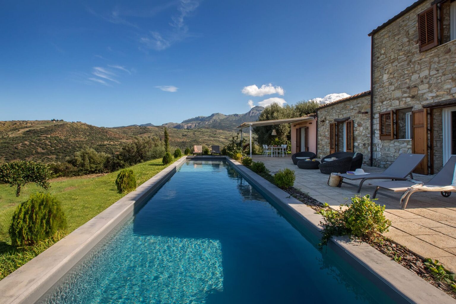 Luxurious stone villa with a modern swimming pool overlooking scenic hills and mountains, surrounded by lush greenery and a clear blue sky. | Only on SopranoVillas
