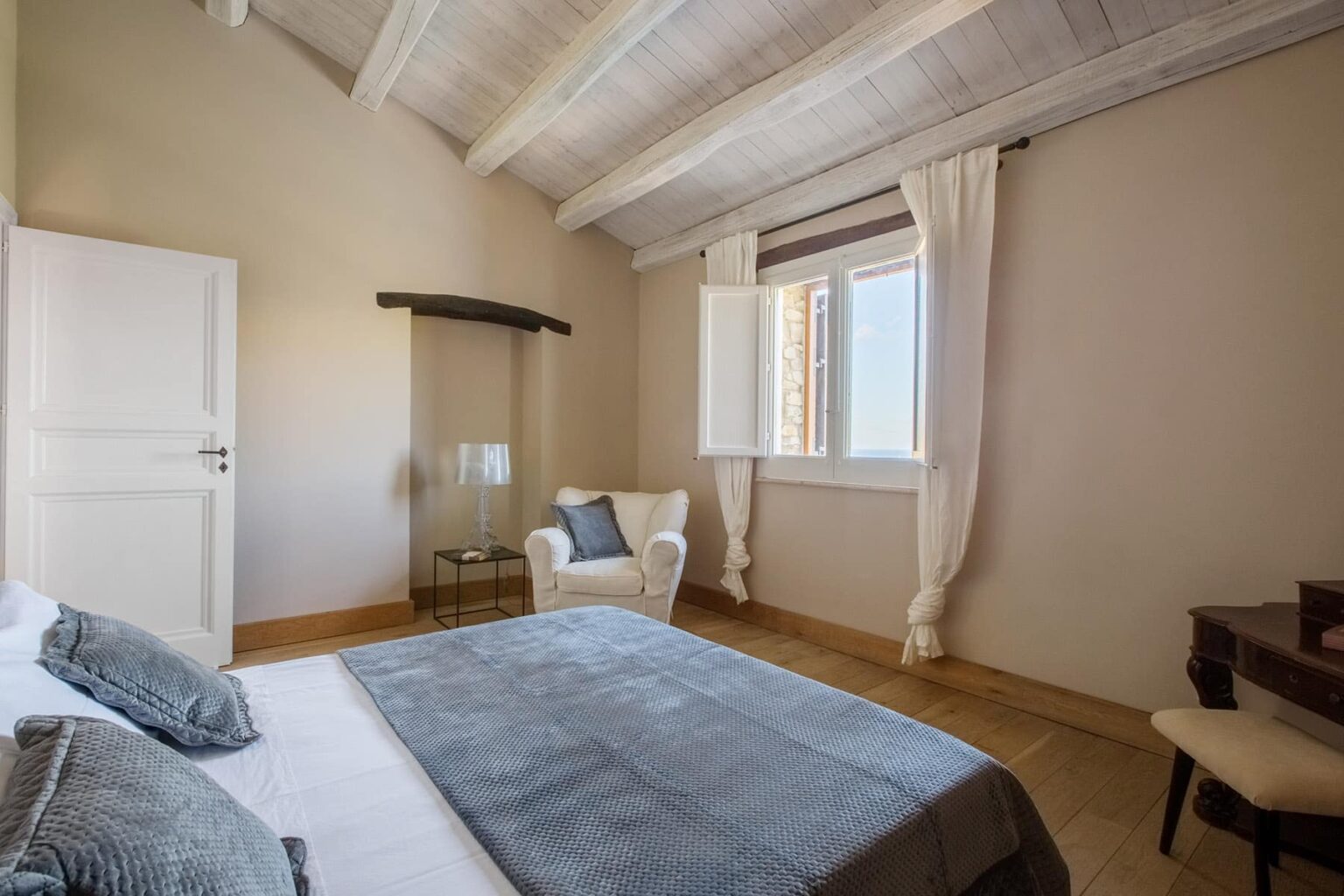 Cozy bedroom featuring a large bed with gray bedding, a comfortable armchair, and large windows with white curtains, all set under a wooden ceiling. | Only on SopranoVillas